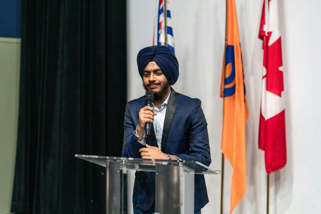 Young Middle Eastern man in turban speaking at a lectern with national flags.