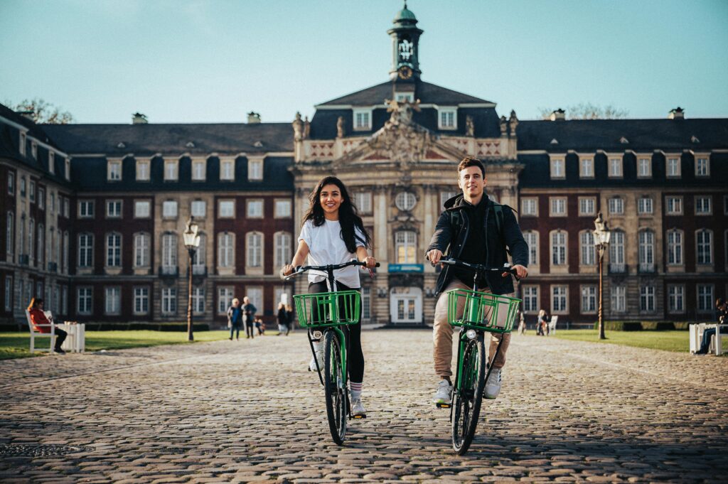 A couple rides bicycles in front of the University of Münster on a sunny day.