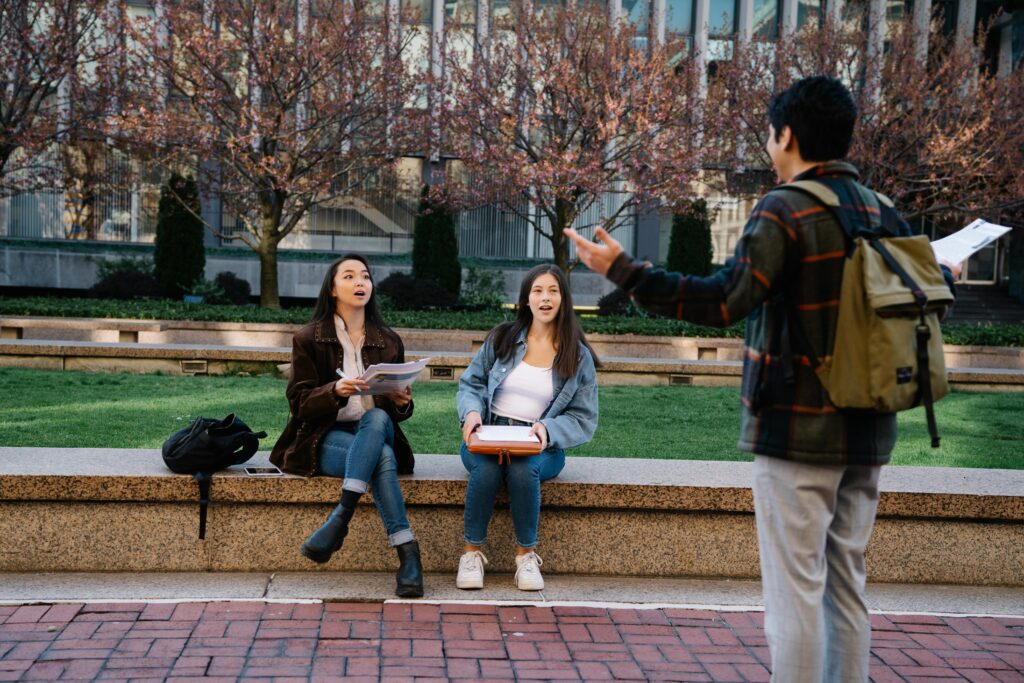 Two female students seated on a bench engage with a standing male student in an outdoor campus setting.