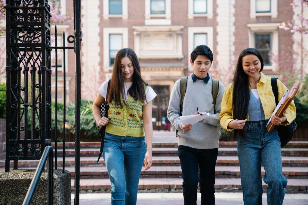 Three smiling students walking together on a university campus, holding notebooks and backpacks.