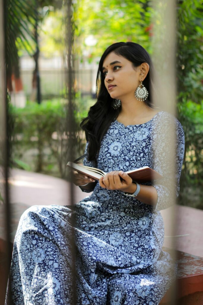 A young woman in a blue dress reading a book outdoors in a garden setting.