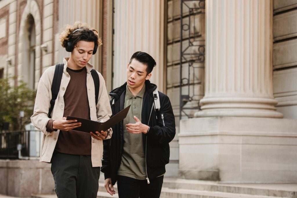 Two young men discussing studies while walking on a university campus outdoors.